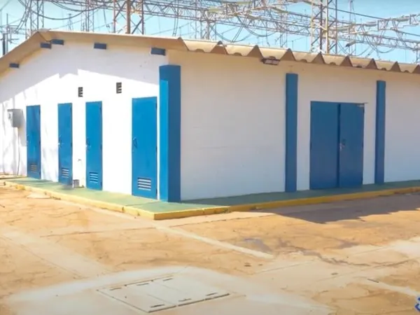 Casa de mando blanca con puertas azules en el patio de una subestación, con estructuras de alta tensión al fondo