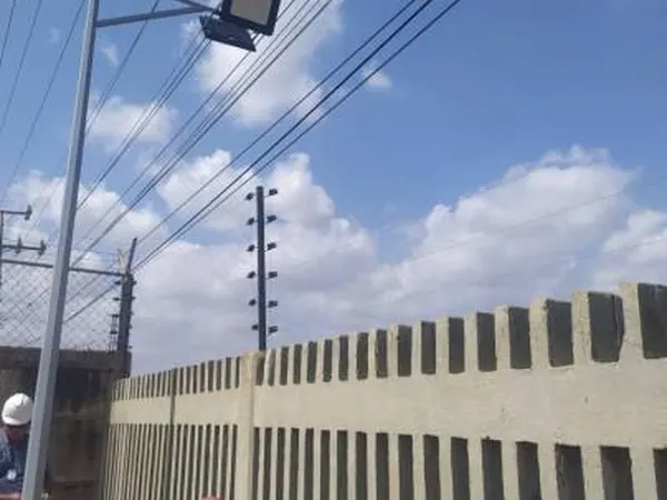Reflector LED en un poste junto a un muro perimetral con cerco eléctrico, bajo un cielo con nubes